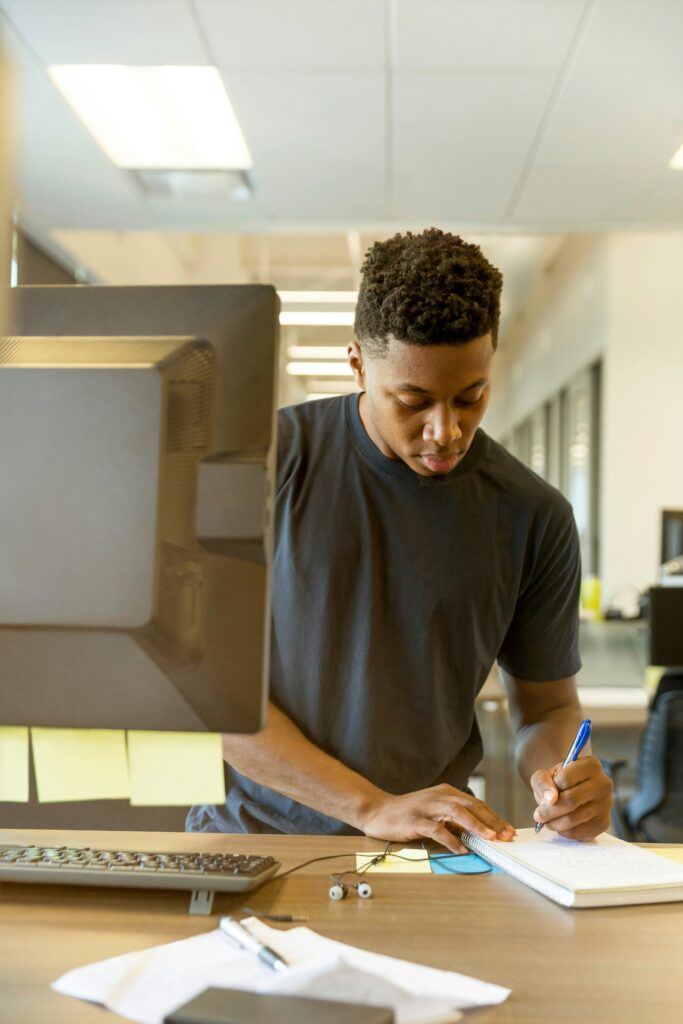 A man writing down information standing next to a computer. 